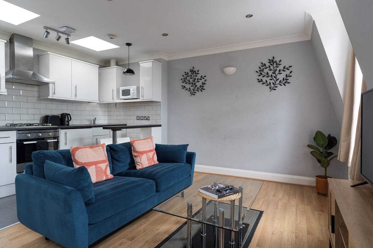 A modern kitchen with white cabinets, a microwave, and a stainless steel range hood. There is a blue sofa with orange and white cushions, a small round table with a magazine, and a potted plant.