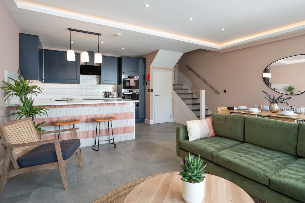 A modern apartment with a kitchen, dining area, and living room. It includes a bar area with stools, a green sofa, and a coffee table with a plant.