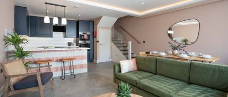 A modern apartment with a kitchen, dining area, and living room. It includes a bar area with stools, a green sofa, and a coffee table with a plant.