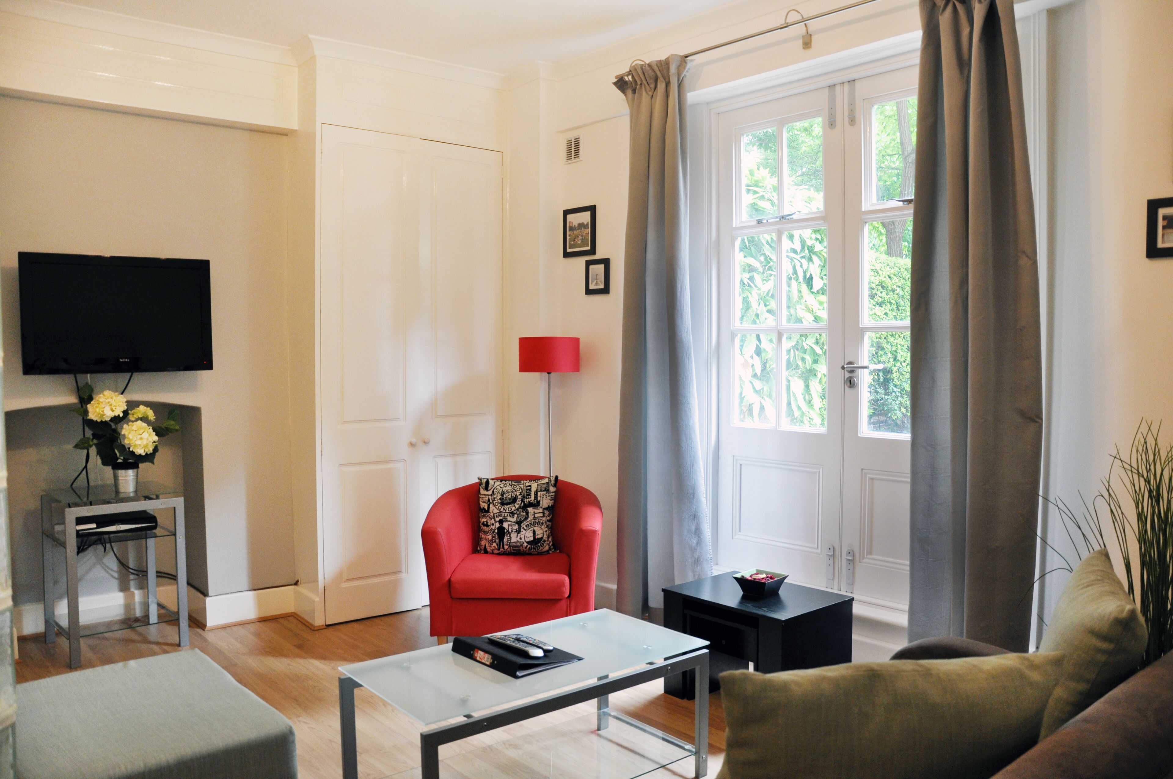 A living room with a red armchair, a glass coffee table, a flat-screen TV mounted on the wall, and a window with curtains.