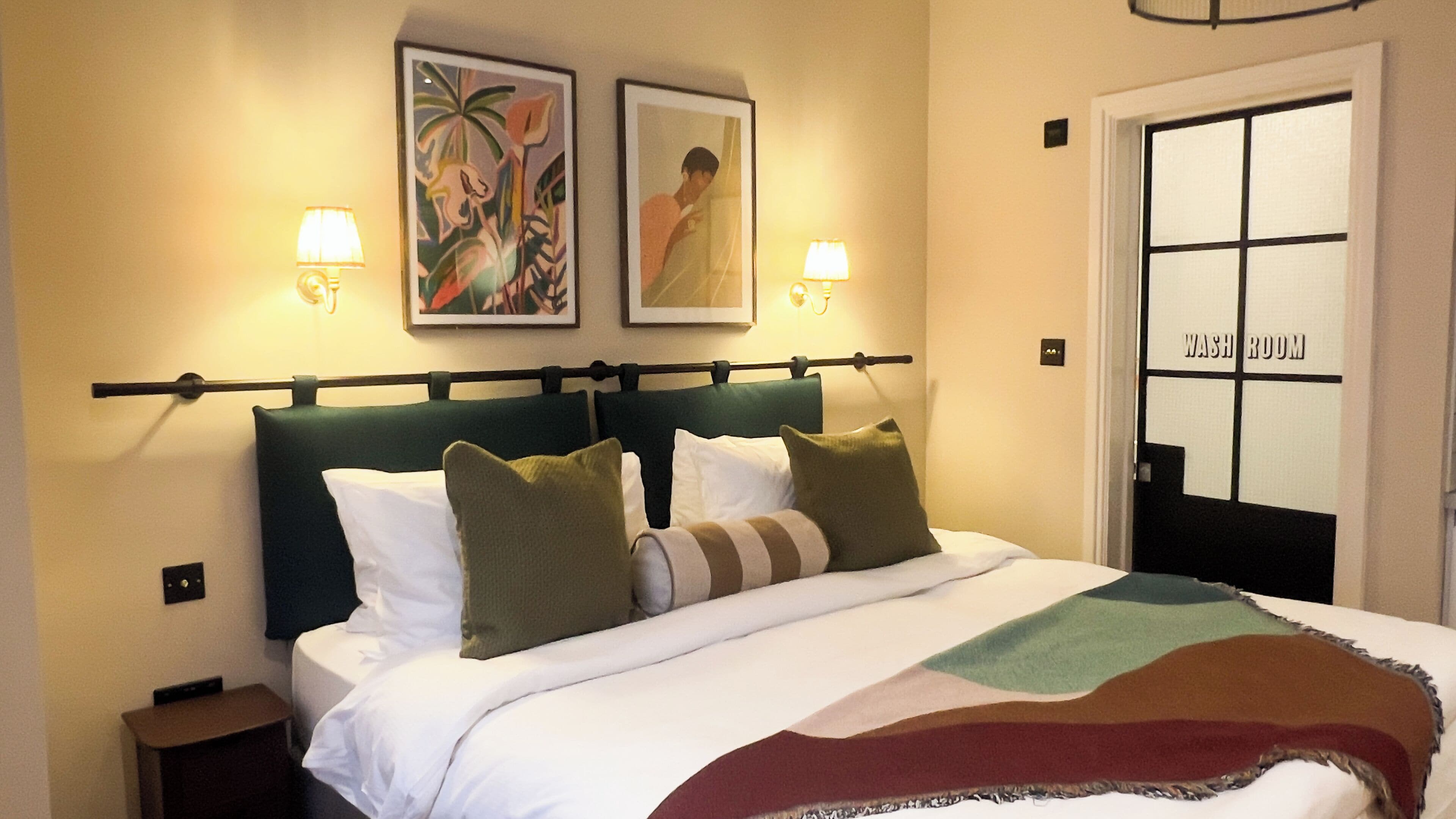 A hotel room with a bed, two wall-mounted lamps, a framed picture, and a door labeled "WASH ROOM".
