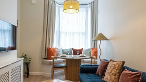 Un salon moderne avec une table ronde en bois, un canapé bleu et un téléviseur posé sur un meuble blanc.