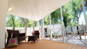 Un espacio al aire libre techado con piso de madera, mobiliario de mimbre y vistas a árboles y un edificio al fondo.
