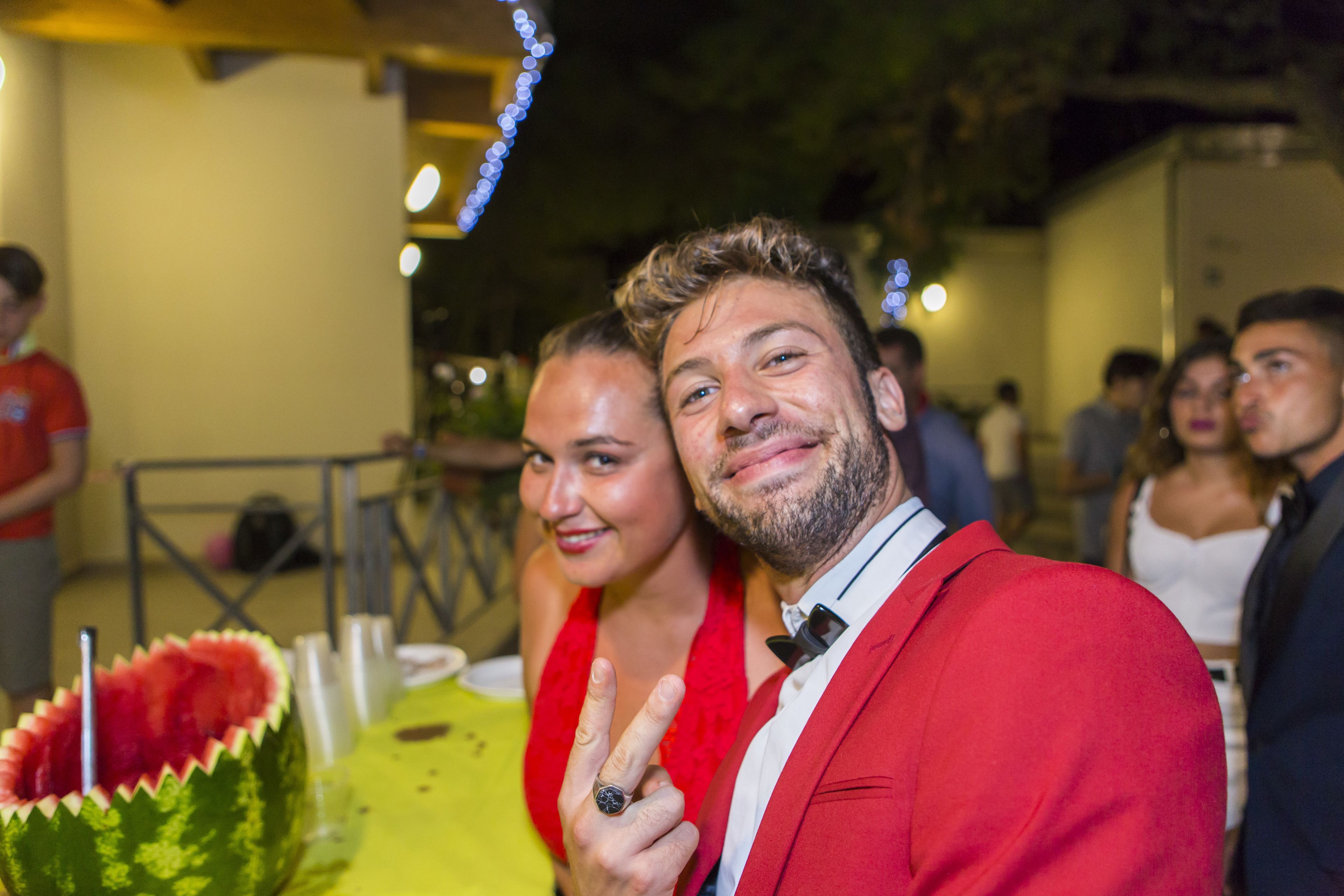 A person in a red suit at a social event with a decorated table and other attendees in the background.