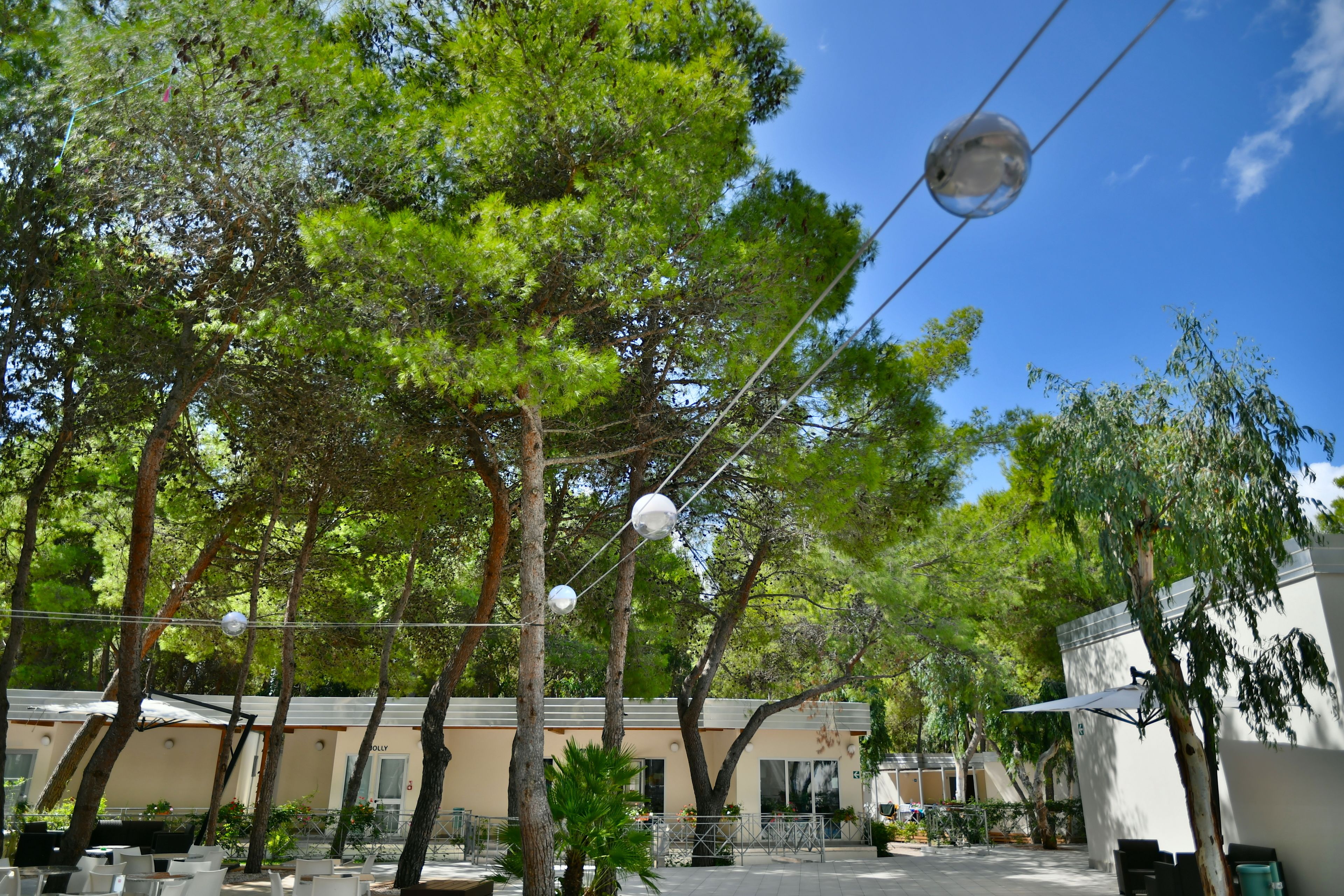 An outdoor seating area with tables and chairs, surrounded by trees and a building with a beige facade.