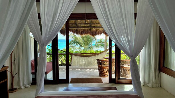 A bedroom with a bed, white curtains, and a view of the ocean and palm trees. - The Beach Tulum Hotel (Tulum)