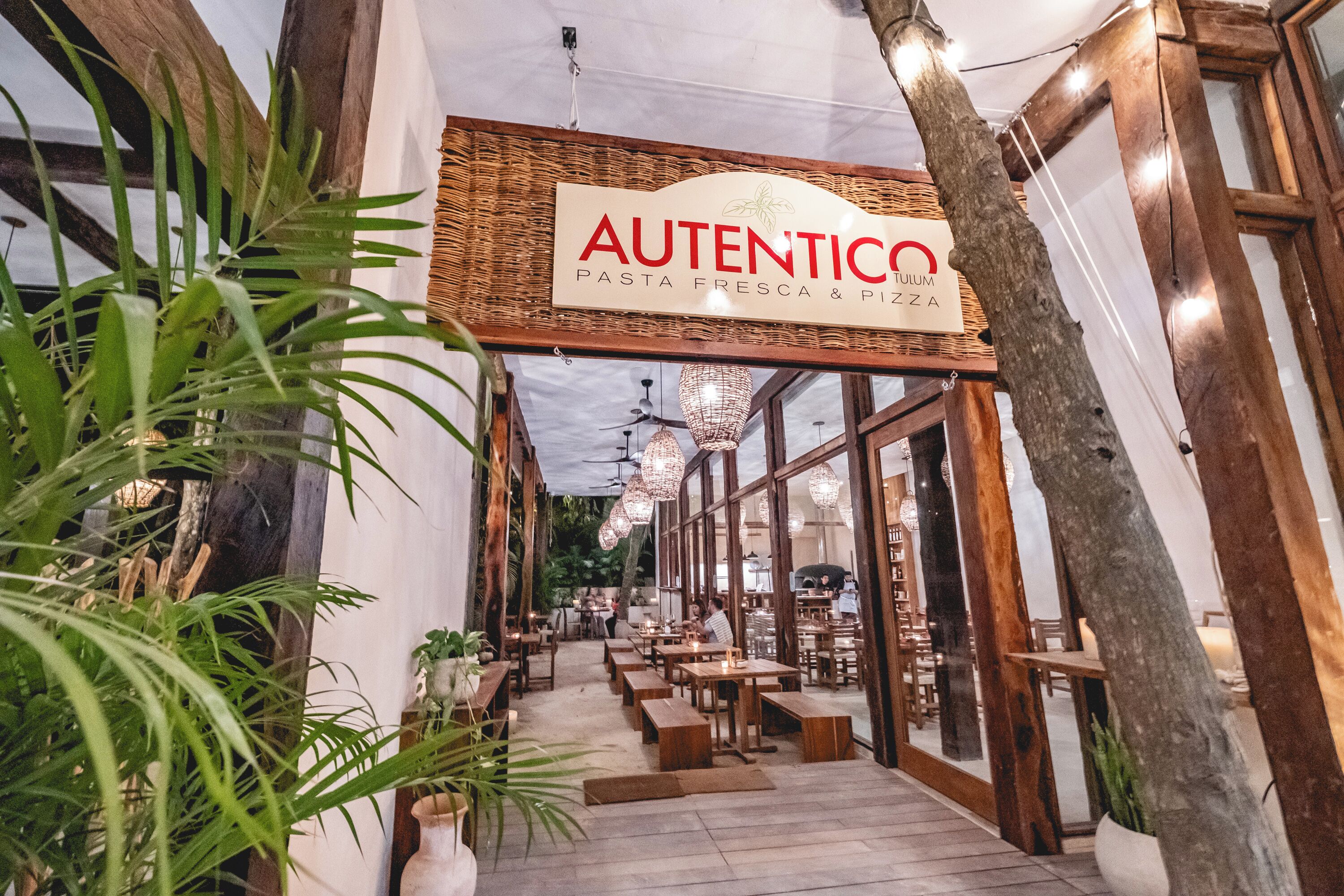Un espace de restauration intérieur avec une enseigne indiquant « Authentico Pasta Fresca & Pizza », des tables en bois, des plantes en pot et des luminaires suspendus.