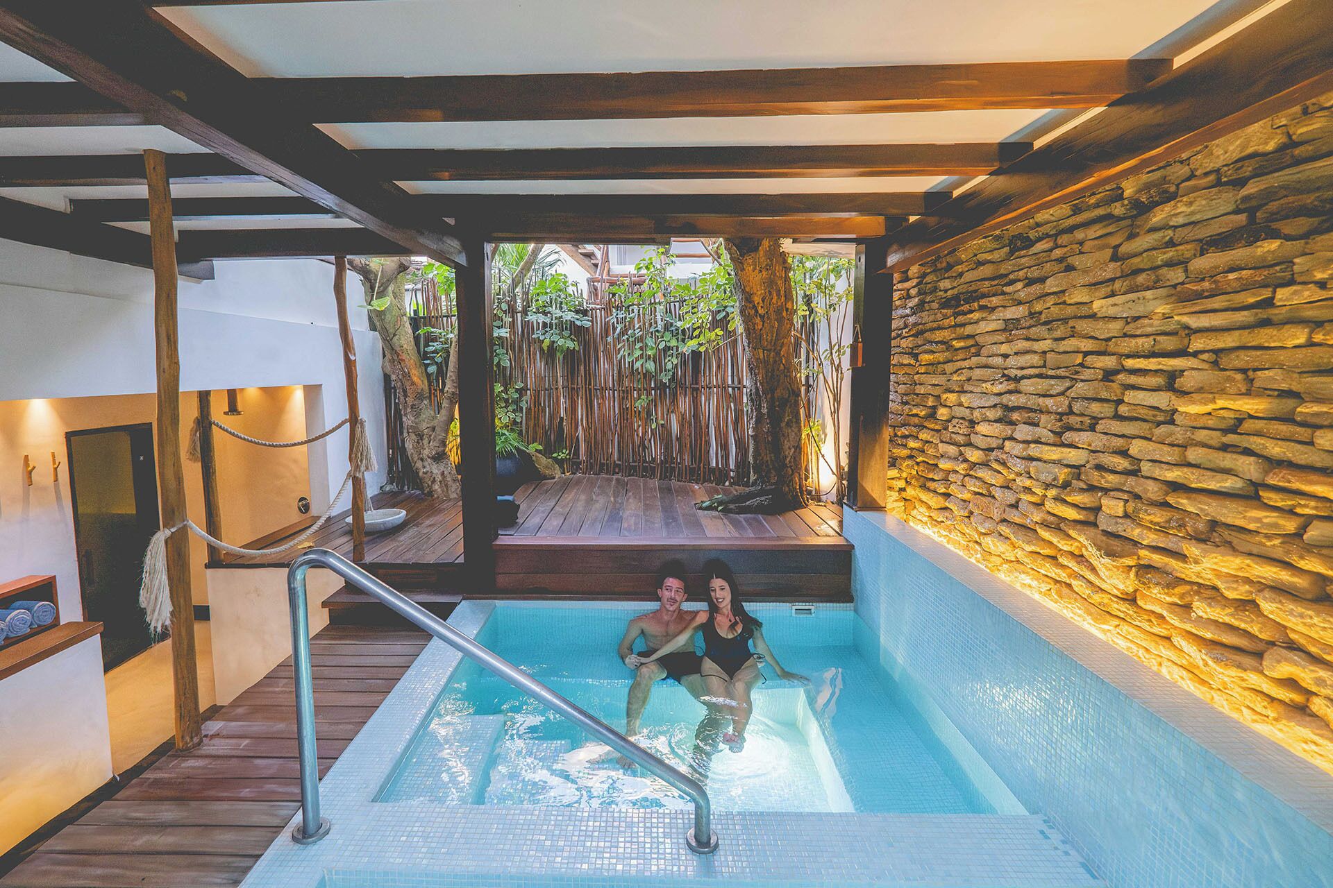 An indoor swimming pool with a wooden deck and stone wall, surrounded by bamboo and plants.