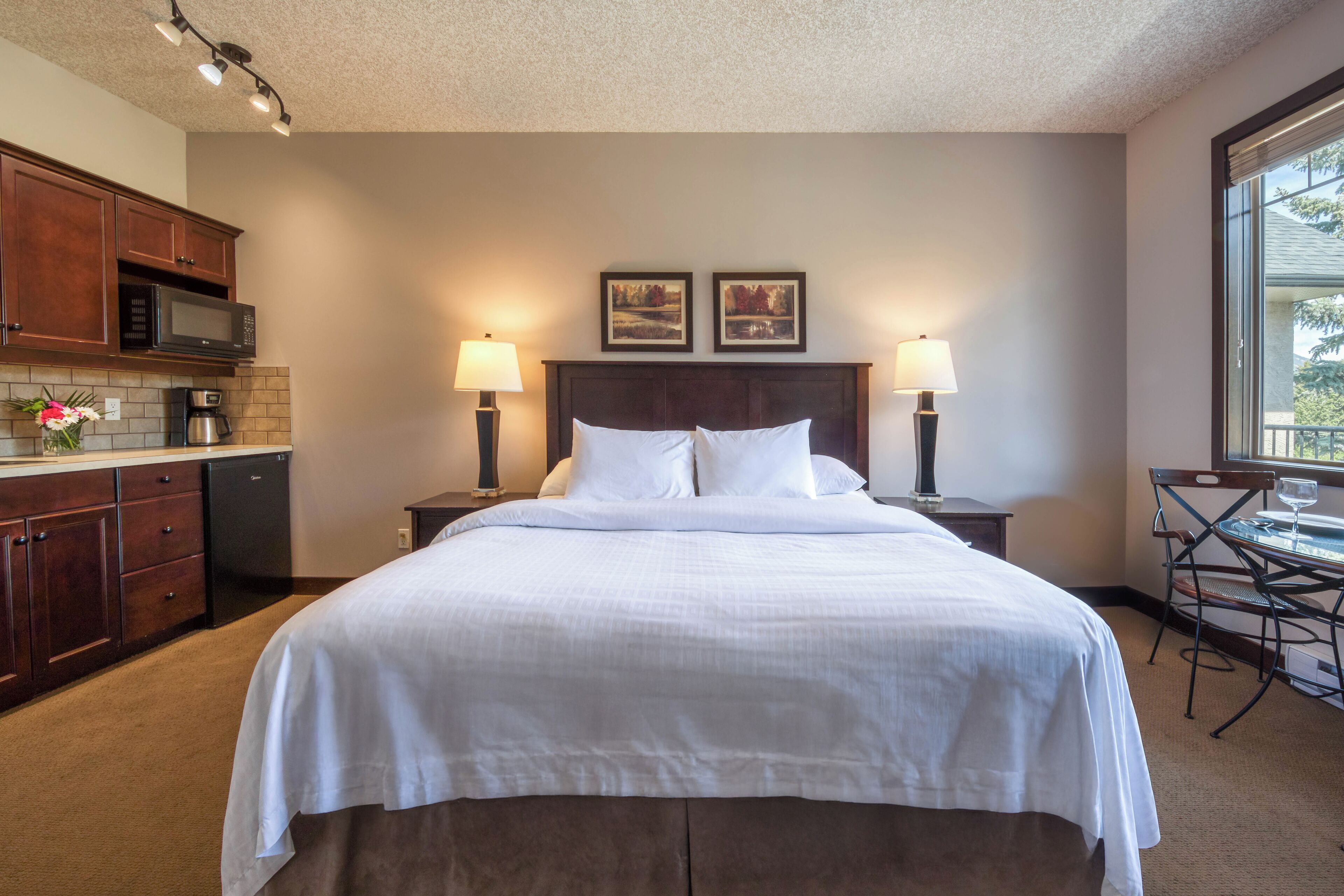 A hotel room with a large bed, two bedside lamps, a small table with a chair, a kitchenette with a microwave, and a coffee maker.