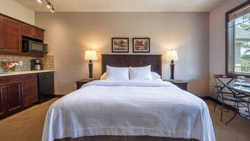 A hotel room with a large bed, two bedside lamps, a small table with a chair, a kitchenette with a microwave, and a coffee maker.