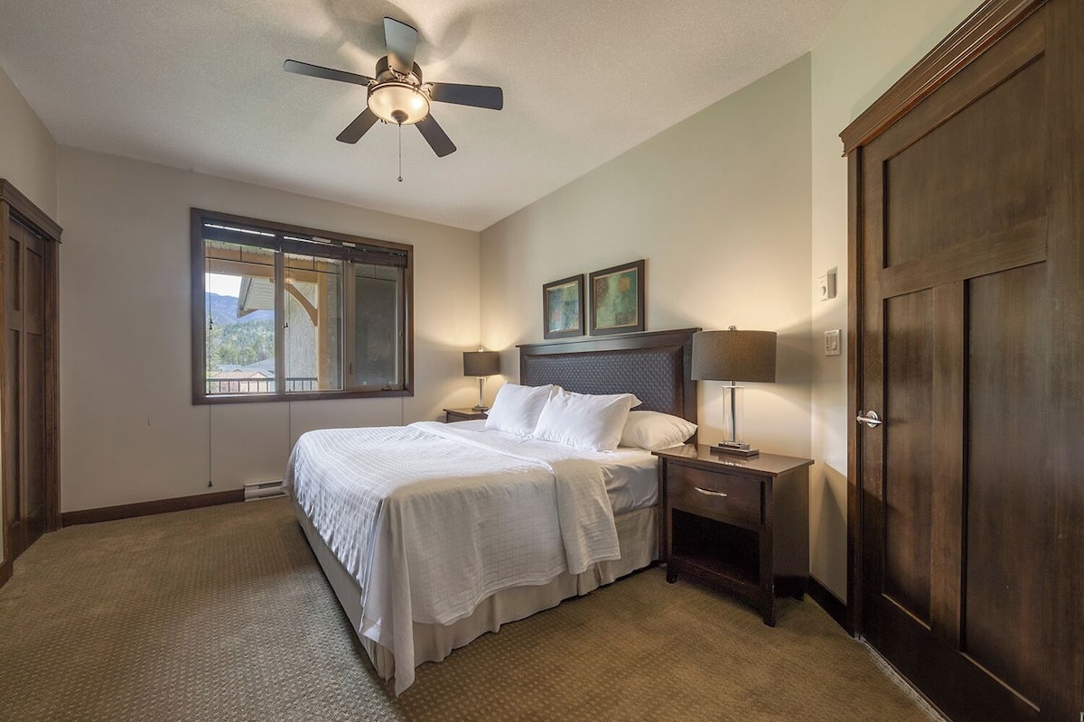A bedroom with a bed, bedside tables, a ceiling fan, a window, and a door.