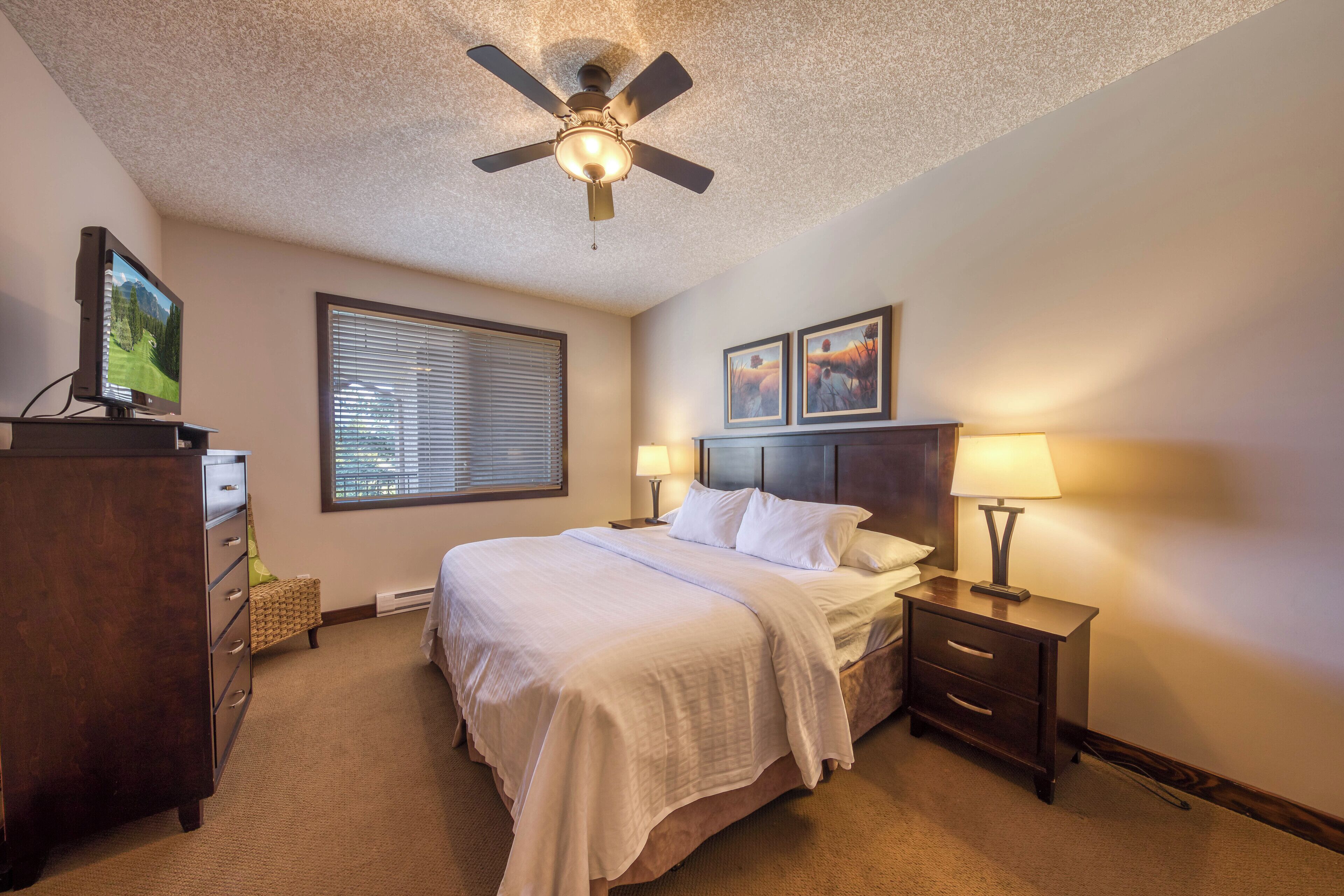 A hotel room with a bed, a ceiling fan, a TV, a window with blinds, two bedside lamps, and a dresser.