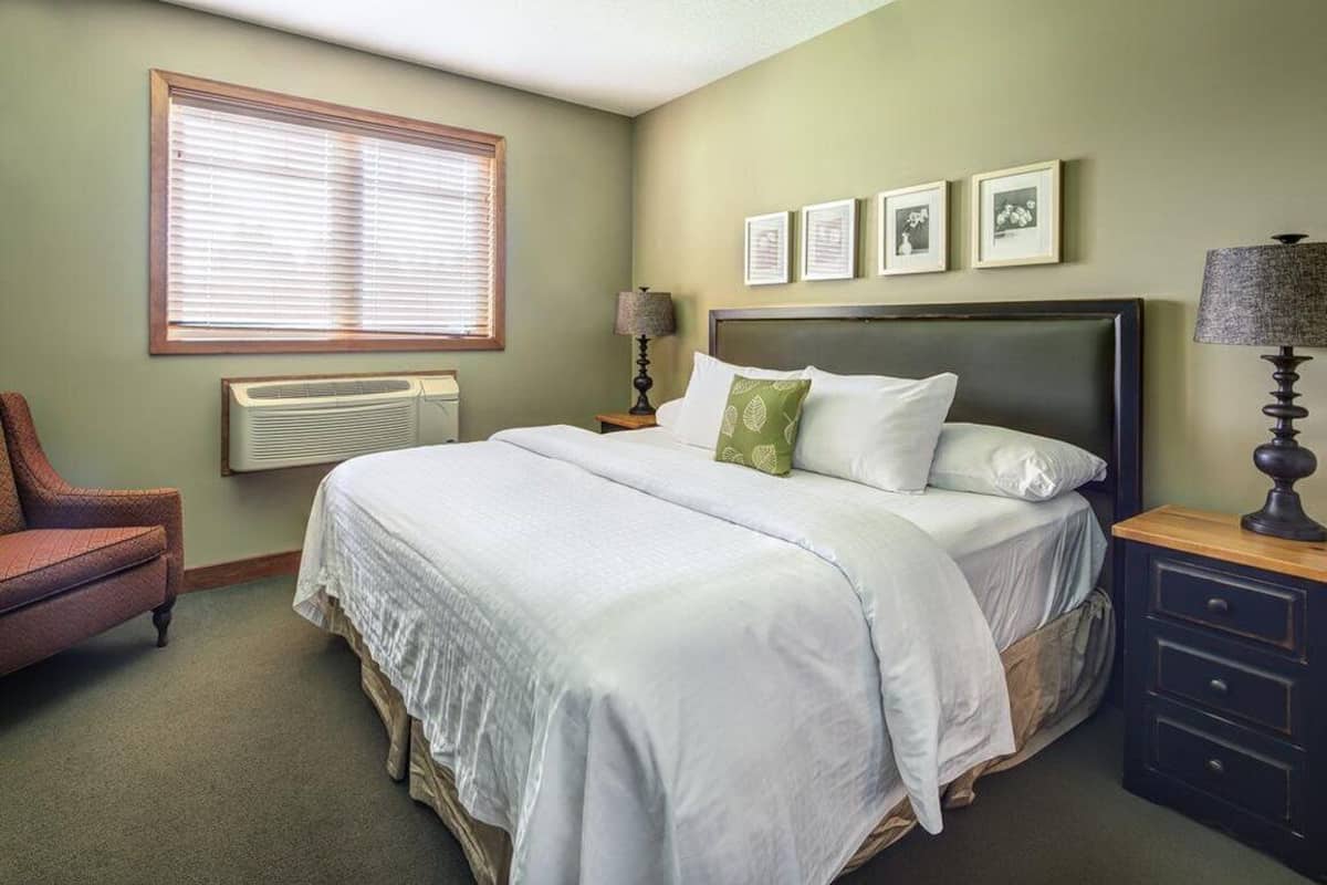 A bedroom with a bed, a chair, a nightstand, a lamp, a window with blinds, and framed pictures on the wall.