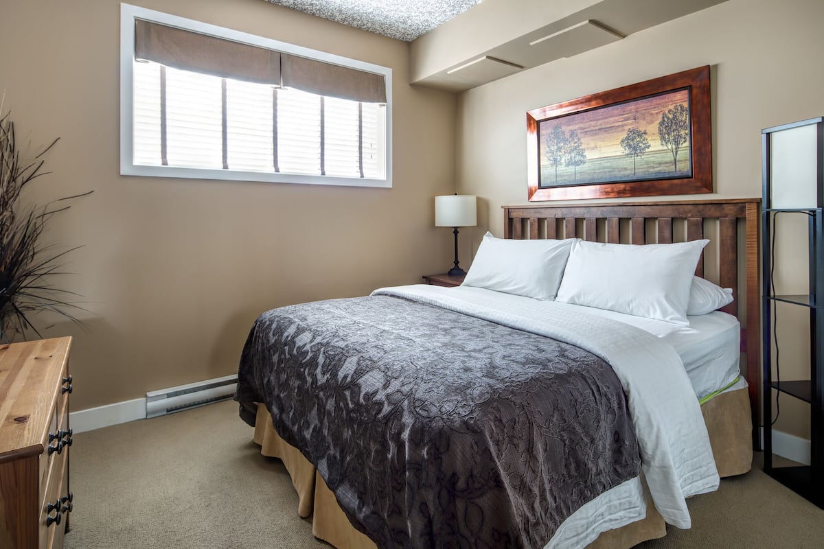 A bedroom with a bed, a nightstand, a lamp, a dresser, a window with blinds, and a painting on the wall.