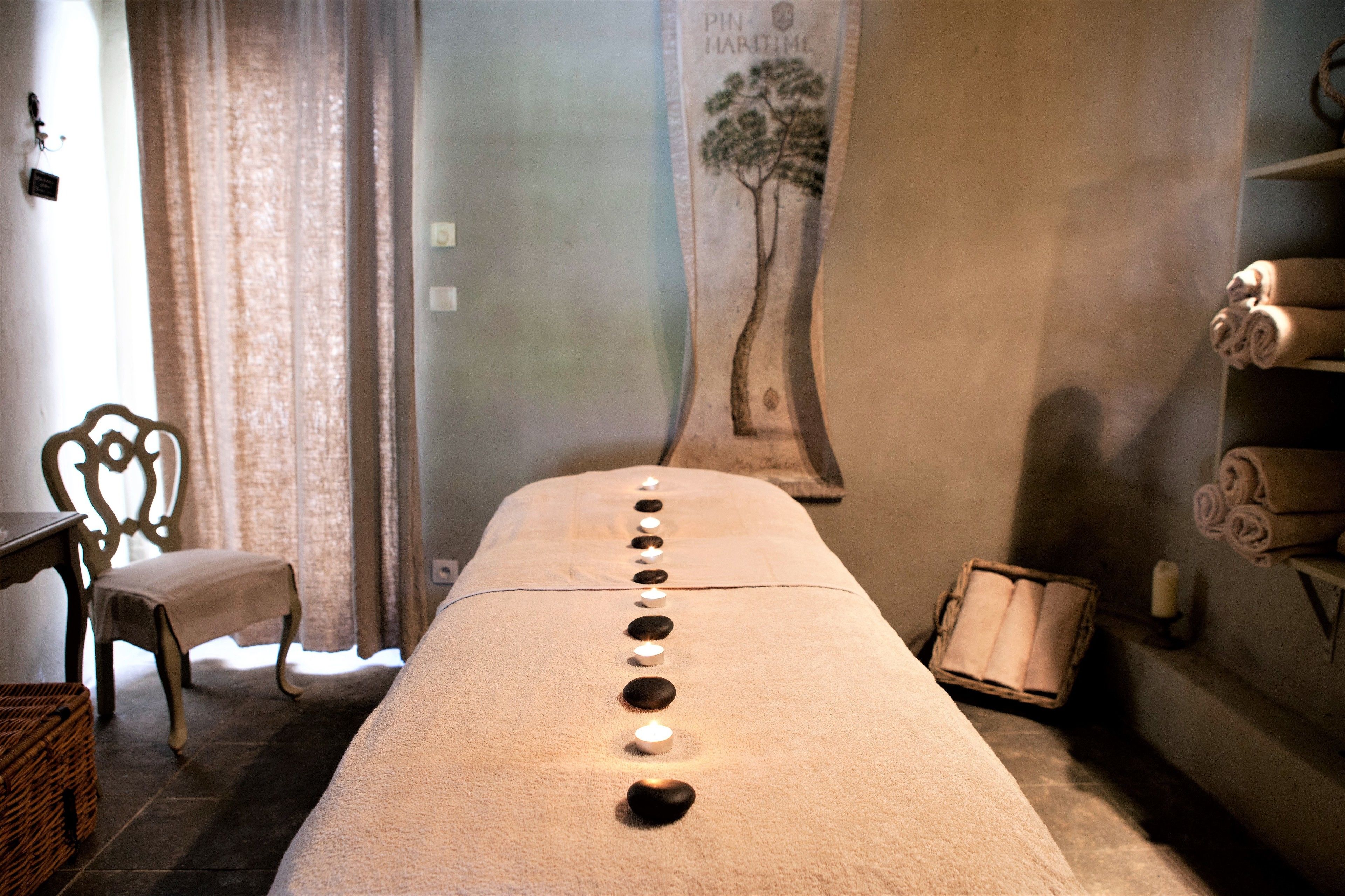 Une pièce comprenant une longue table de massage plate, des bougies, une chaise, un mur orné d’une fresque représentant un arbre et des étagères avec des serviettes.