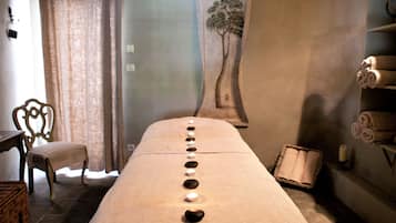 Une pièce comprenant une longue table de massage plate, des bougies, une chaise, un mur orné d’une fresque représentant un arbre et des étagères avec des serviettes.