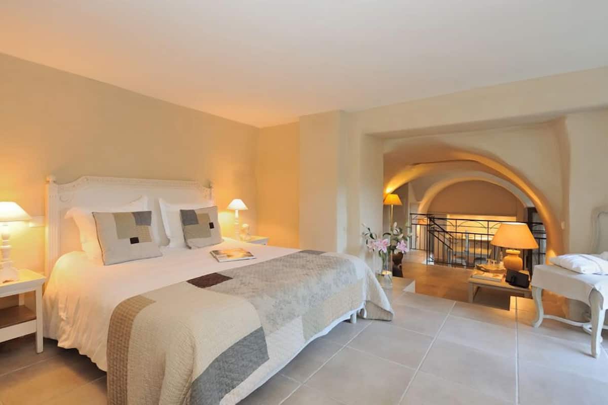 A spacious bedroom with a large bed, two bedside tables, a nightstand, and a chair. There is a staircase leading to an upper level.