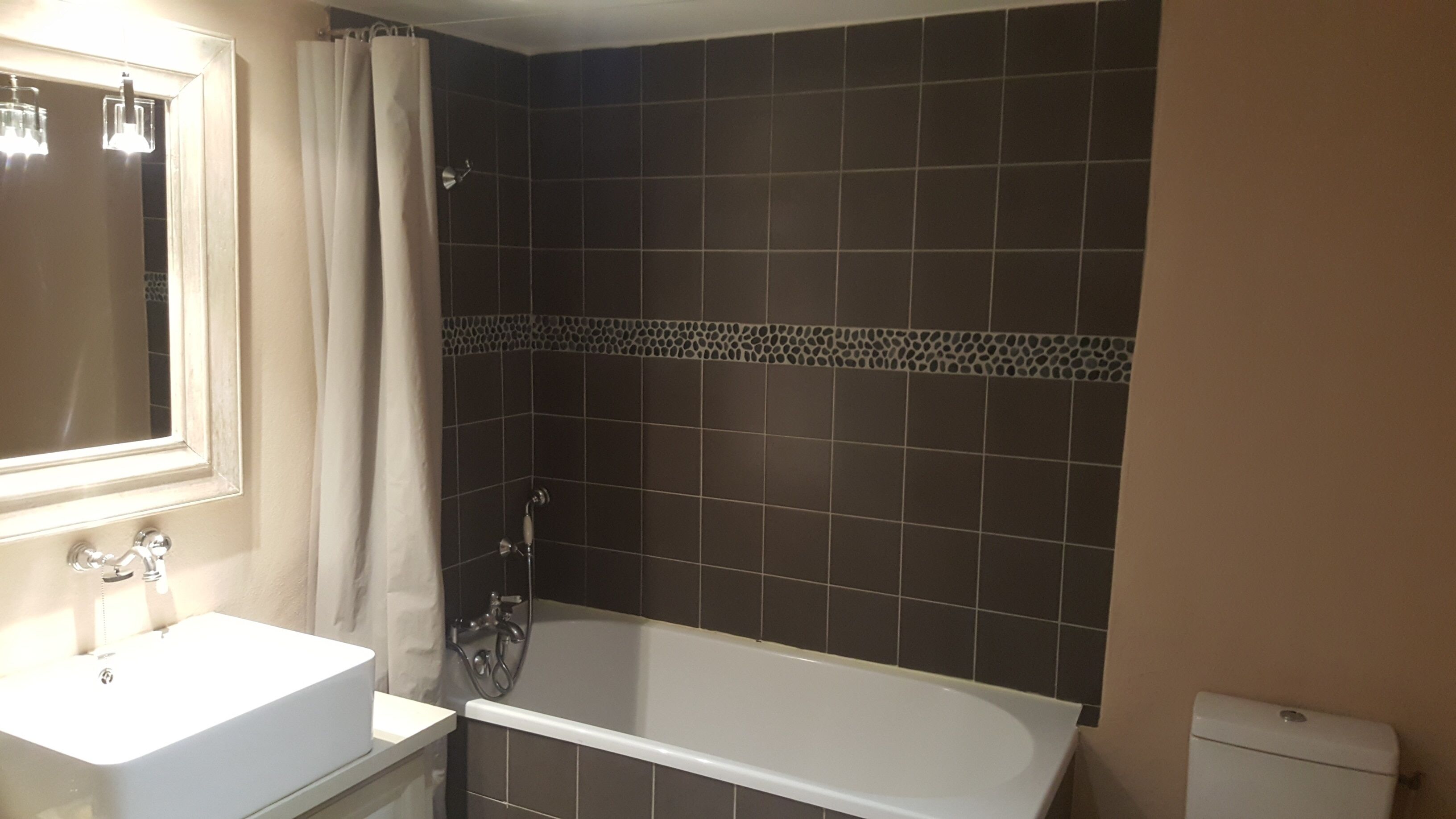 Un baño con lavamanos blanco, espejo, bañera y paredes de baldosas oscuras.