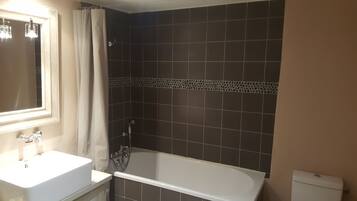 Un baño con lavamanos blanco, espejo, bañera y paredes de baldosas oscuras.