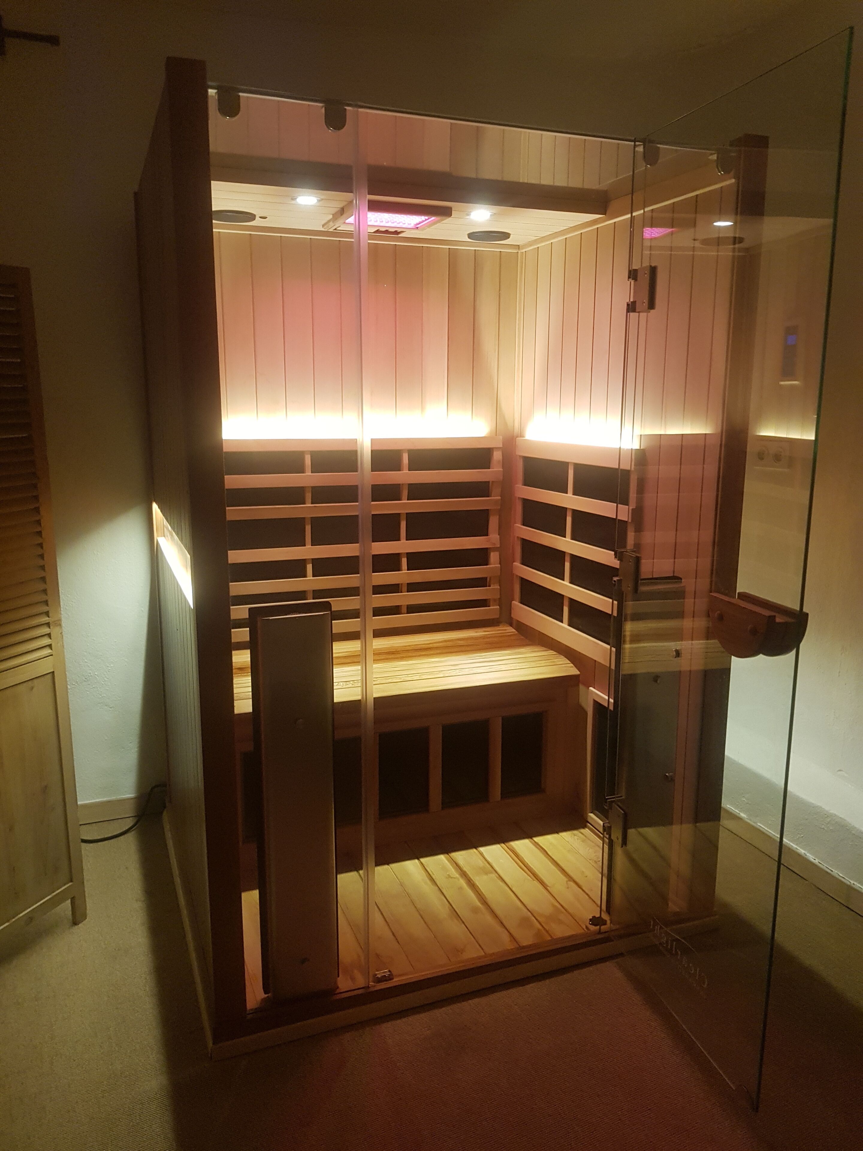 A sauna with wooden benches and glass walls, featuring overhead lighting and a visible ceiling vent.
