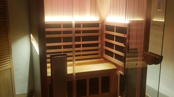 A sauna with wooden benches and glass walls, featuring overhead lighting and a visible ceiling vent.