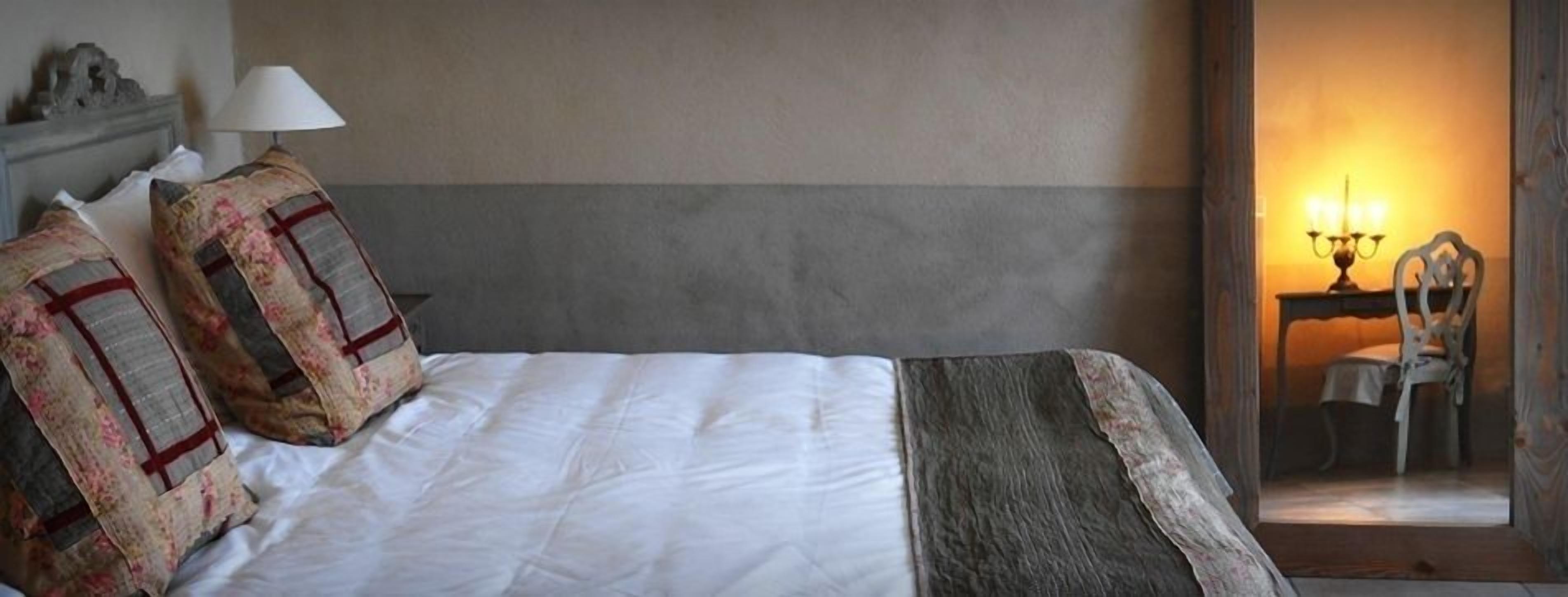 Una habitación de hotel con cama, almohadas, una lámpara, una silla y una mesa con candelabro.