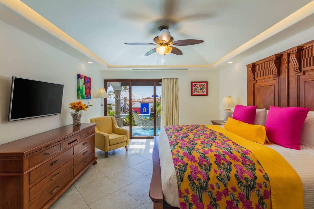 Habitación de hotel con cama, televisión, cómoda de madera, una silla y vistas a una piscina y edificios.