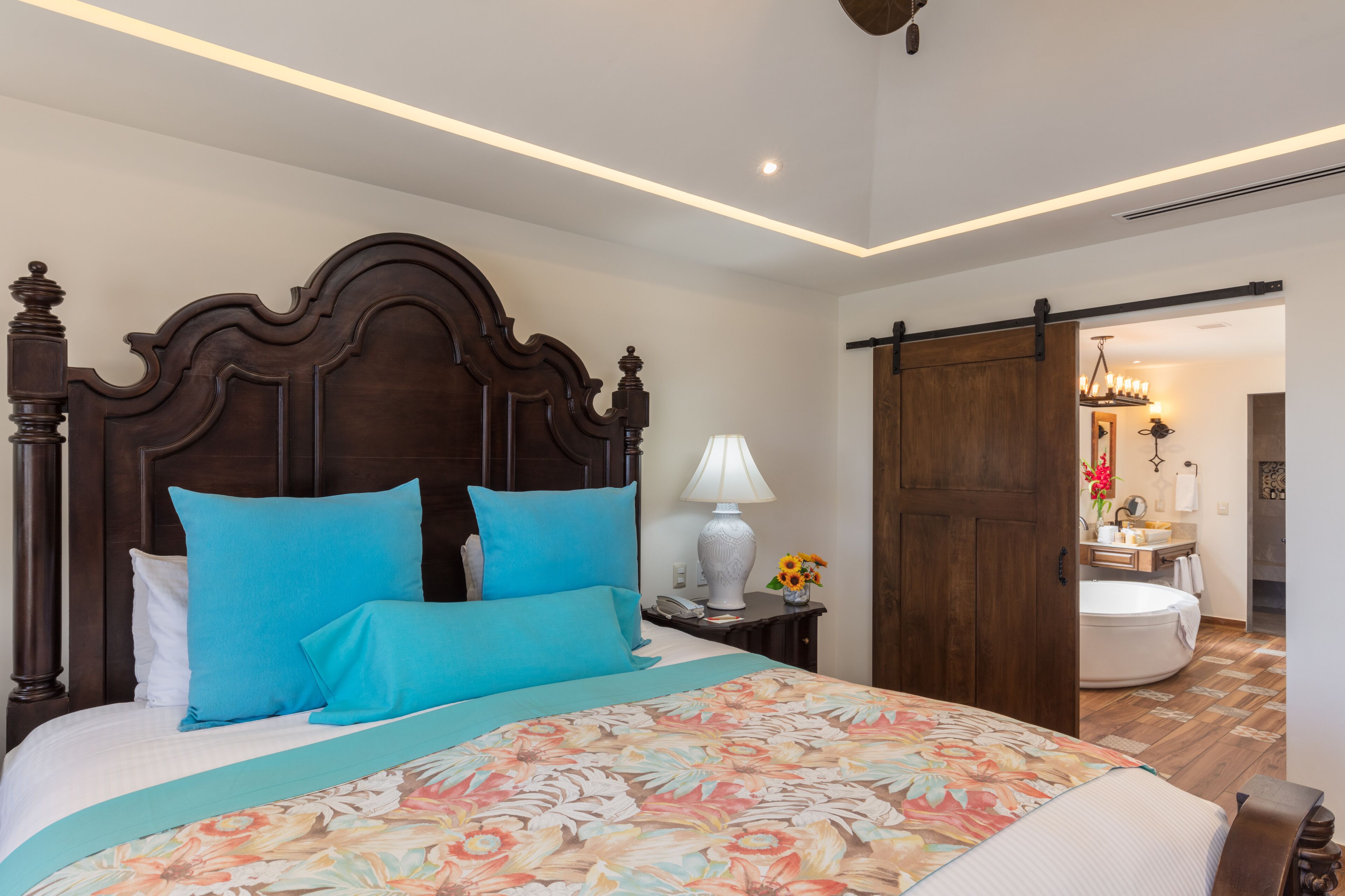 Un dormitorio con una cama grande, cabecera de madera, mesita de noche con lámpara, y un baño visible a través de una puerta abierta.