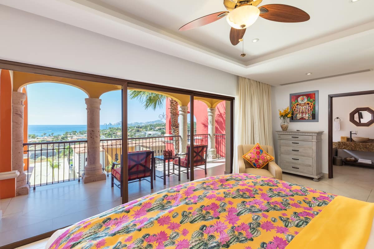 Un dormitorio con balcón, una cama con colcha estampada, ventilador de techo y vista al océano.