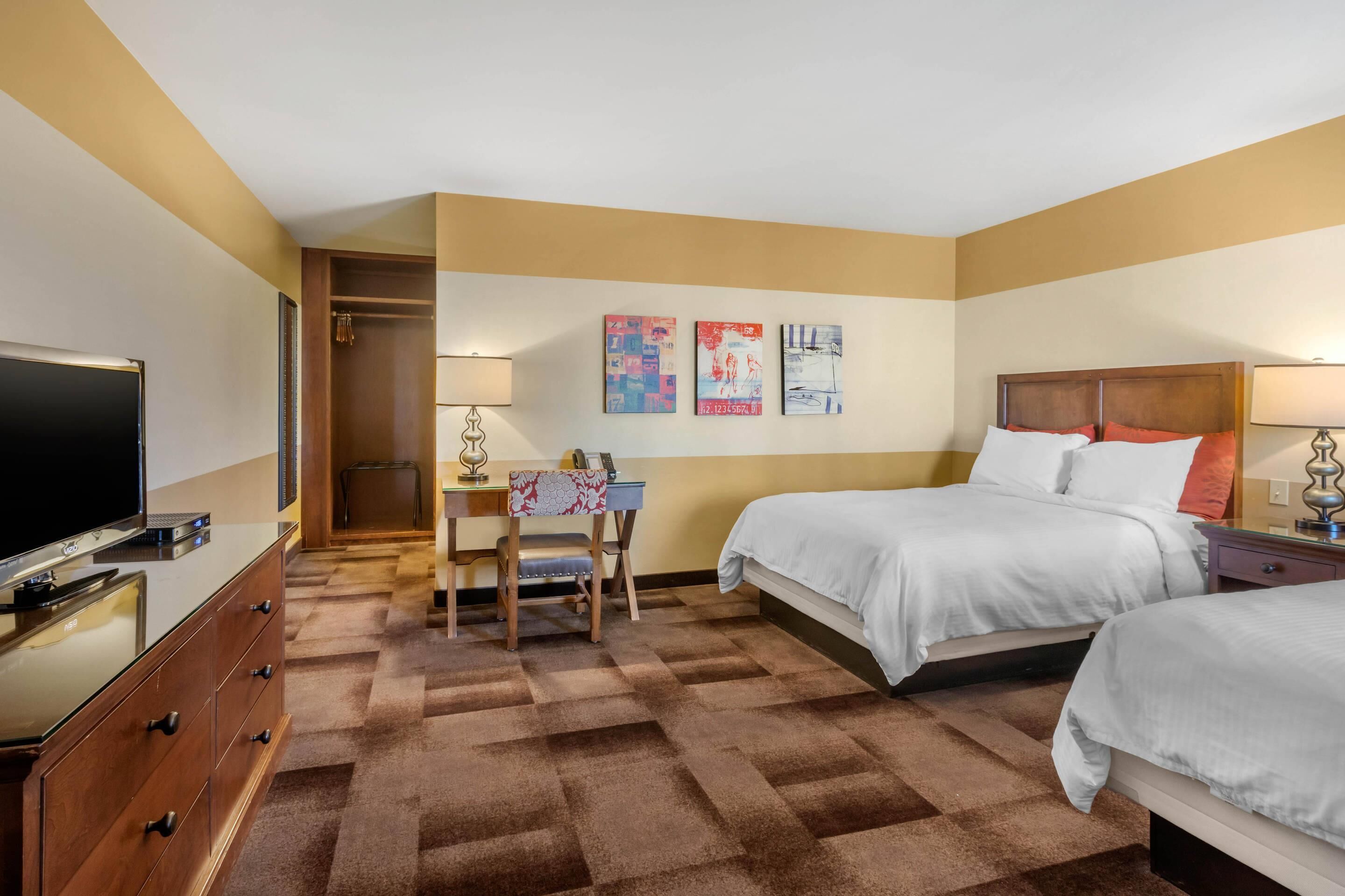 Quarto de hotel com duas camas, uma escrivaninha com cadeira, televisão e obras de arte na parede.