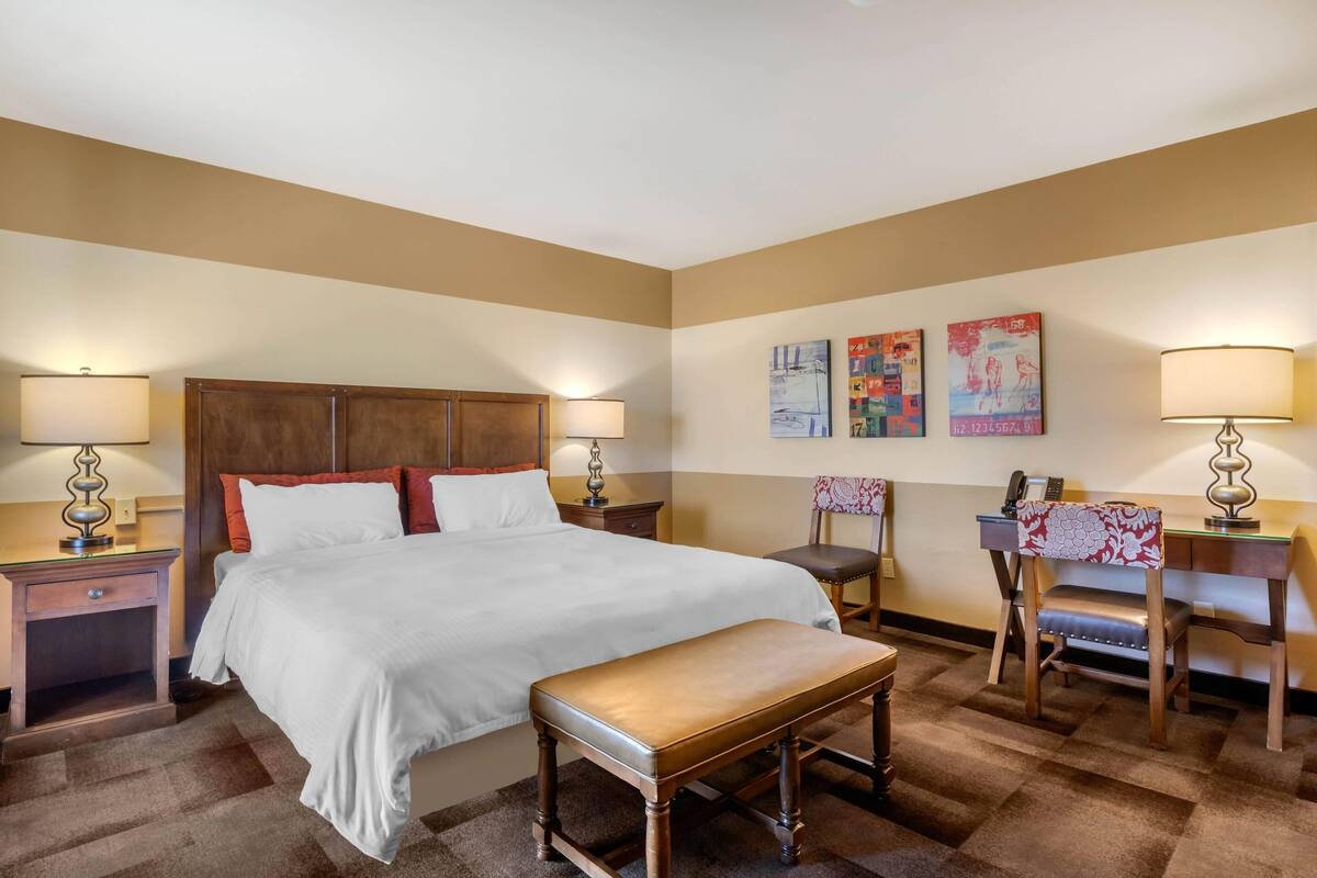 A hotel room with a bed, bedside tables, a desk, a chair, and artwork on the wall.