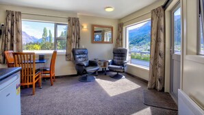 Ein Zimmer mit Esstisch, zwei Stühlen, einem kleinen Tisch mit Lampe und Bergblick durch große Fenster.