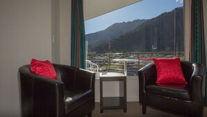 Ein Zimmer mit zwei schwarzen Ledersesseln, jeder mit einem roten Kissen, und einem kleinen Tisch. Ein großes Fenster bietet Blick auf eine gebirgige Landschaft und ein Gewässer.