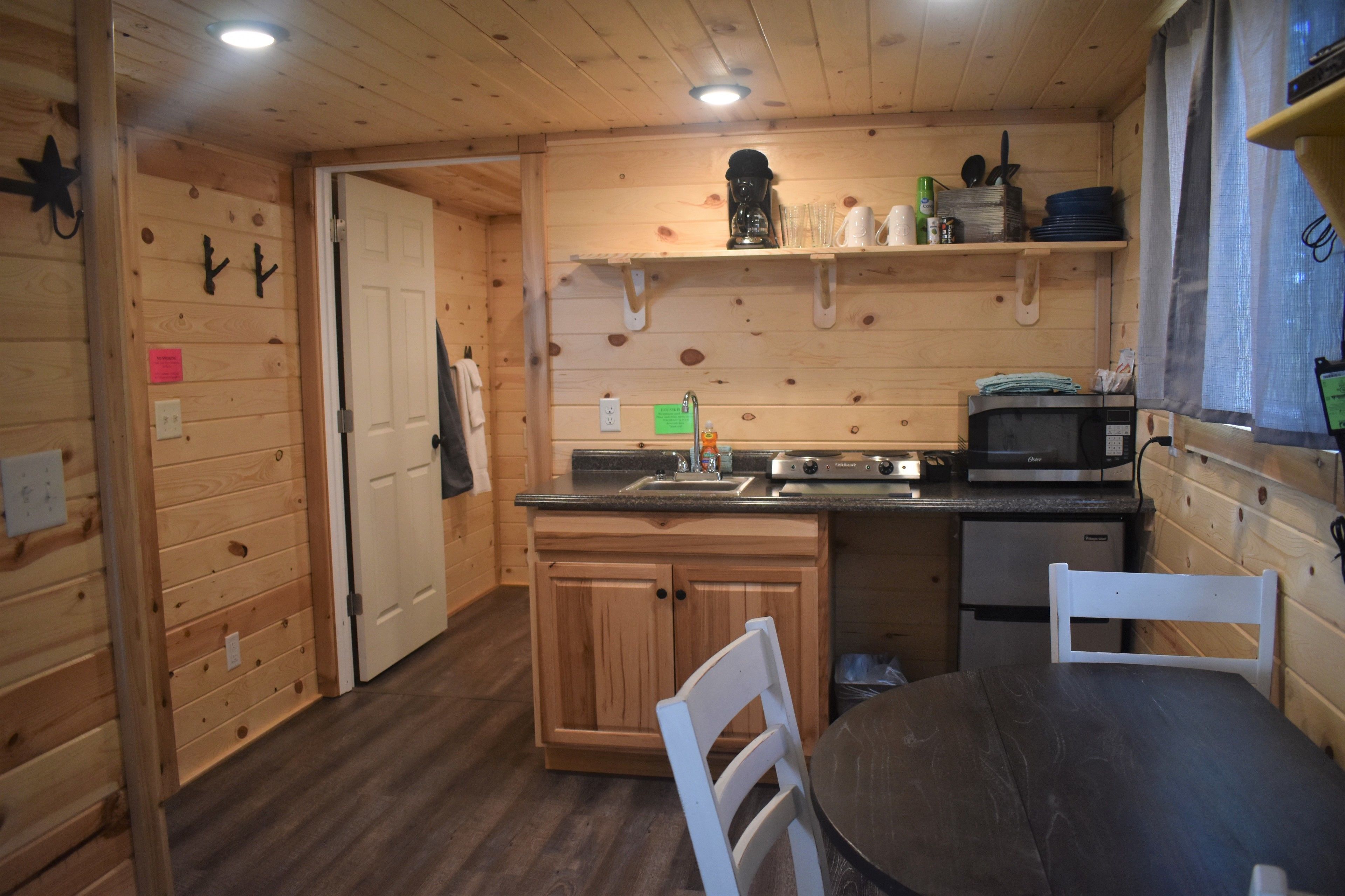 Interior de una cabaña de madera con una zona de cocina que incluye estufa, microondas y pileta. Hay muebles de madera, una mesa de comedor con sillas y una ventana con cortinas.