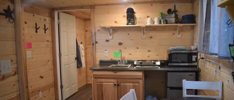 Interior de una cabaña de madera con una zona de cocina que incluye estufa, microondas y pileta. Hay muebles de madera, una mesa de comedor con sillas y una ventana con cortinas.