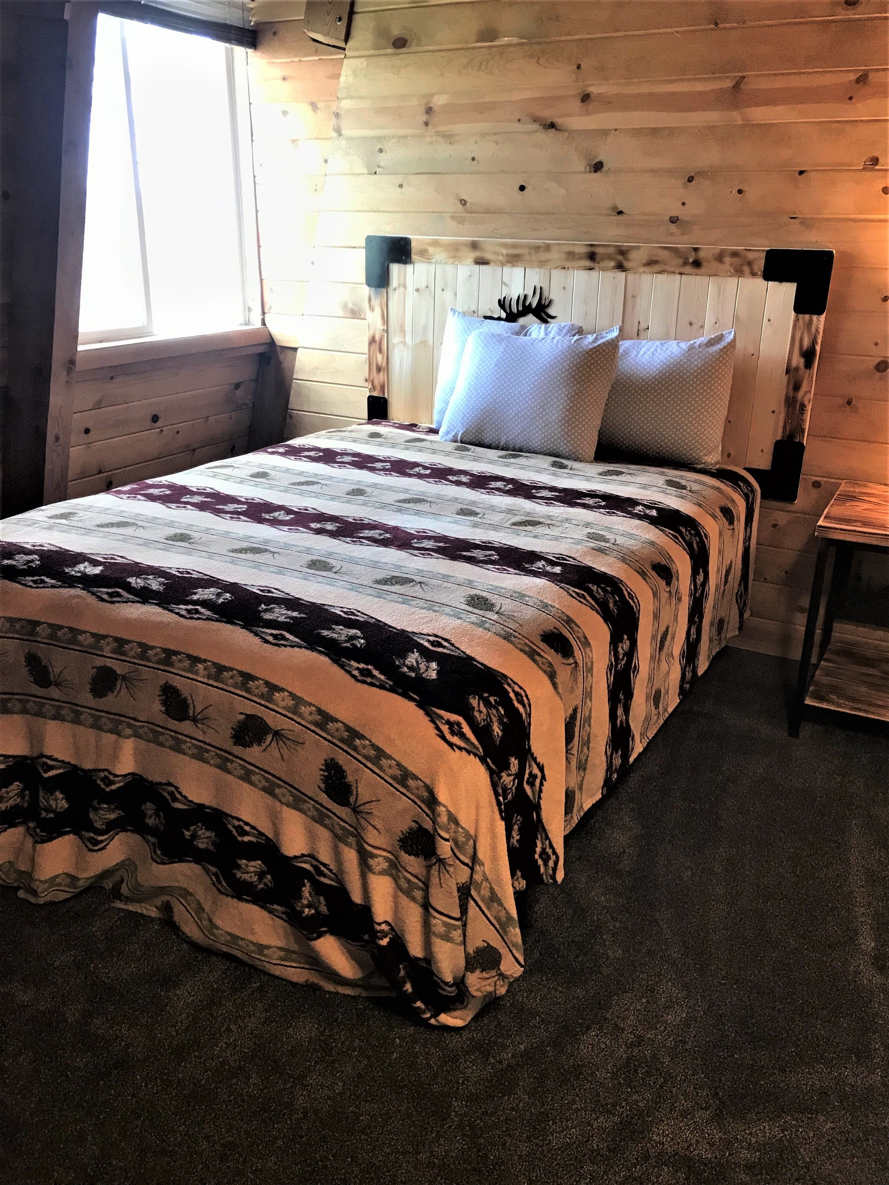 Un dormitorio de cabaña de madera con una cama, dos almohadas, una mesita de noche y una ventana.