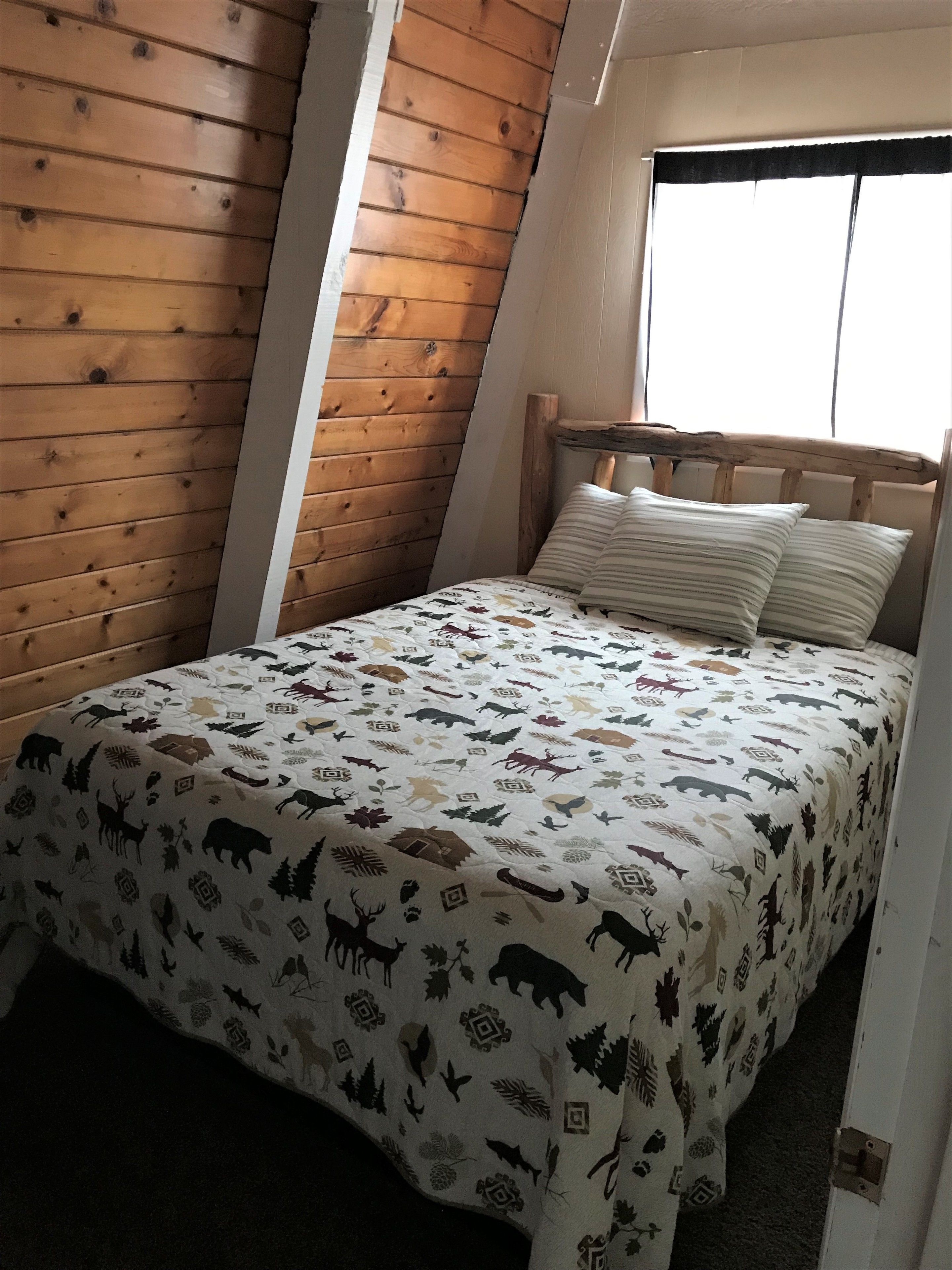 Una camera da letto con soffitto spiovente, pareti in legno, un letto con copripiumino decorato e una finestra con tenda a balza nera.