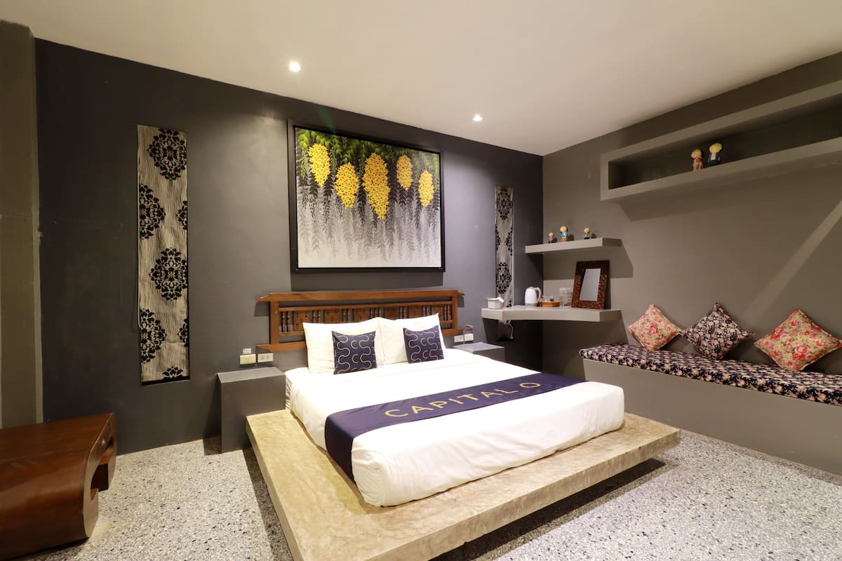 A modern bedroom with a large bed, a headboard, a bedside table, a nightstand, a bench with cushions, a shelf with decorative items, and a wall with a large floral painting.