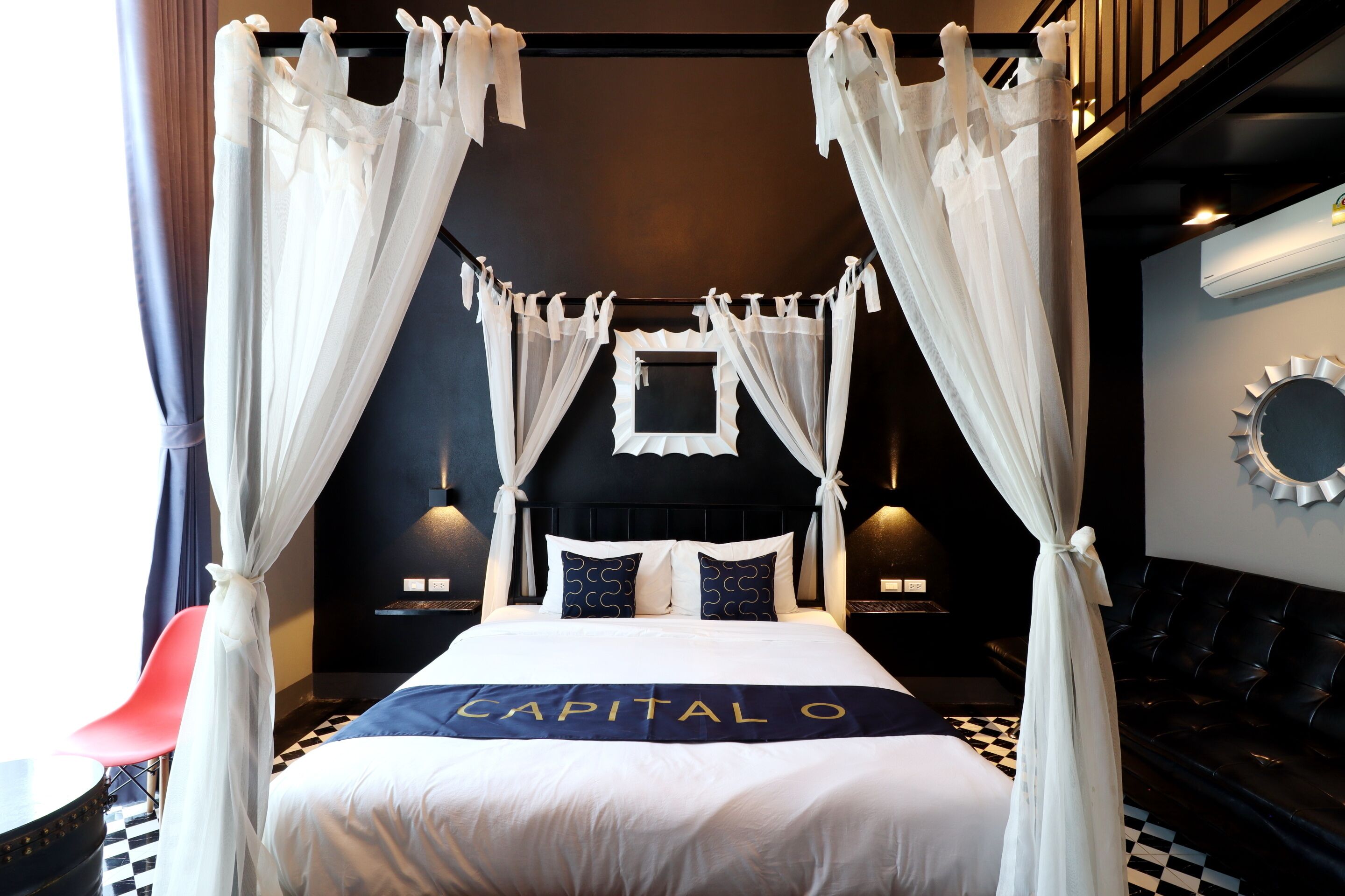 Una cama con dosel y cortinas, un suelo a cuadros blanco y negro, un espejo colgado en la pared y una almohada con la letra 'CAPITAL O'.
