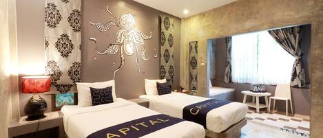 Une chambre d’hôtel avec deux lits portant l’inscription « CAPITAL », une fresque murale décorative, une petite table avec une chaise et une fenêtre avec des rideaux.
