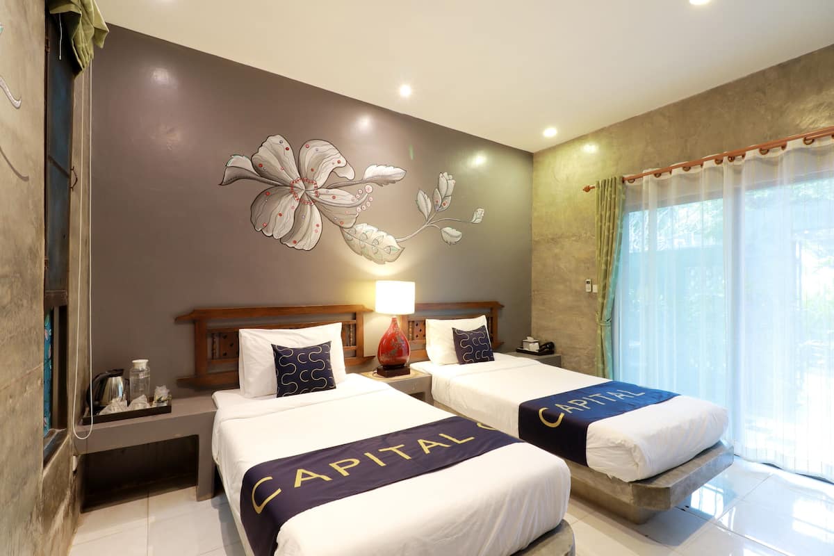 A hotel room with two beds, a floral wall mural, a nightstand with a lamp, and a view of the outdoors through a window with curtains.