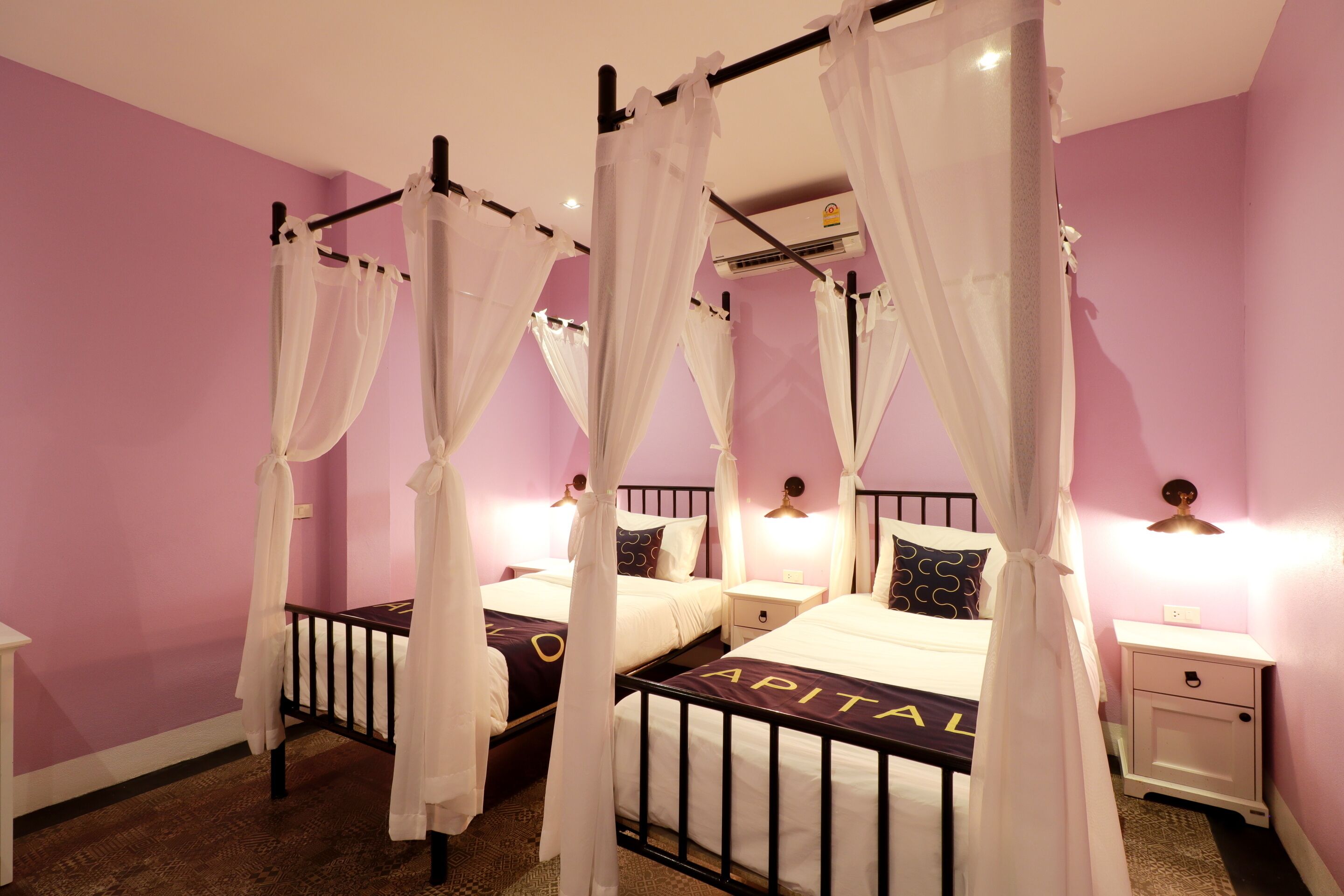 A room with four beds, each with a canopy bed frame and white bedding. There are bedside tables with lamps and a small drawer. The walls are painted pink, and there is an air conditioning unit on the wall.
