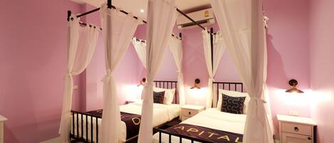 A room with four beds, each with a canopy bed frame and white bedding. There are bedside tables with lamps and a small drawer. The walls are painted pink, and there is an air conditioning unit on the wall.