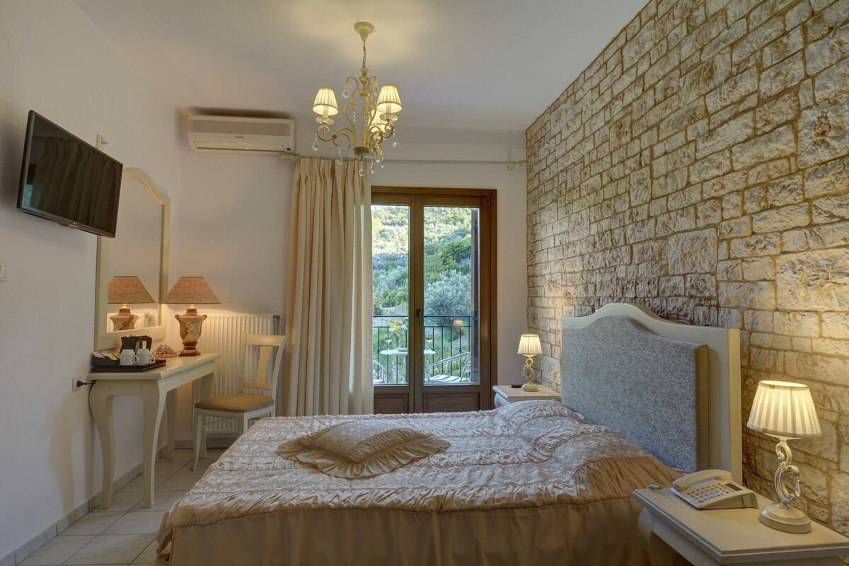 A hotel room with a bed, a desk with a lamp, a chair, a TV, and a balcony with a view of greenery.