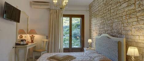 A hotel room with a bed, a desk with a lamp, a chair, a TV, and a balcony with a view of greenery.