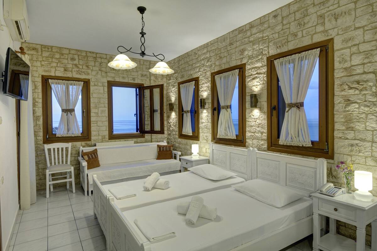 A hotel room with a stone wall, two beds, a chair, a TV, and a view of the sea.