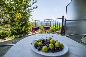 Une table sur laquelle se trouvent une assiette de raisins et deux verres de vin rouge, le tout placé sur un balcon d’où l’on peut admirer la verdure et une clôture.