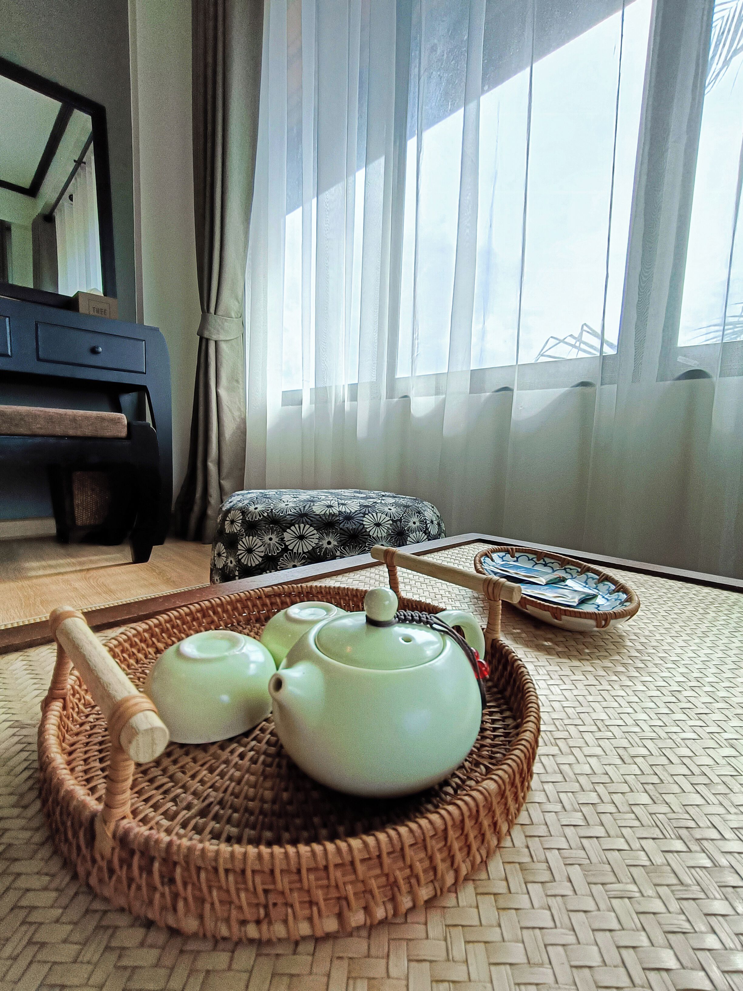 A room with a wicker tray holding teapots and a tea brush, a patterned cushion, and a small table with a tray.
