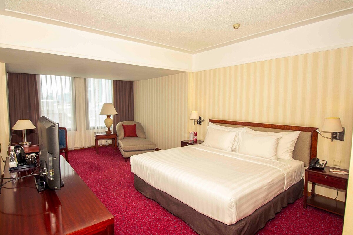 A hotel room with a large bed, a desk with a television, a chair, and a desk lamp.