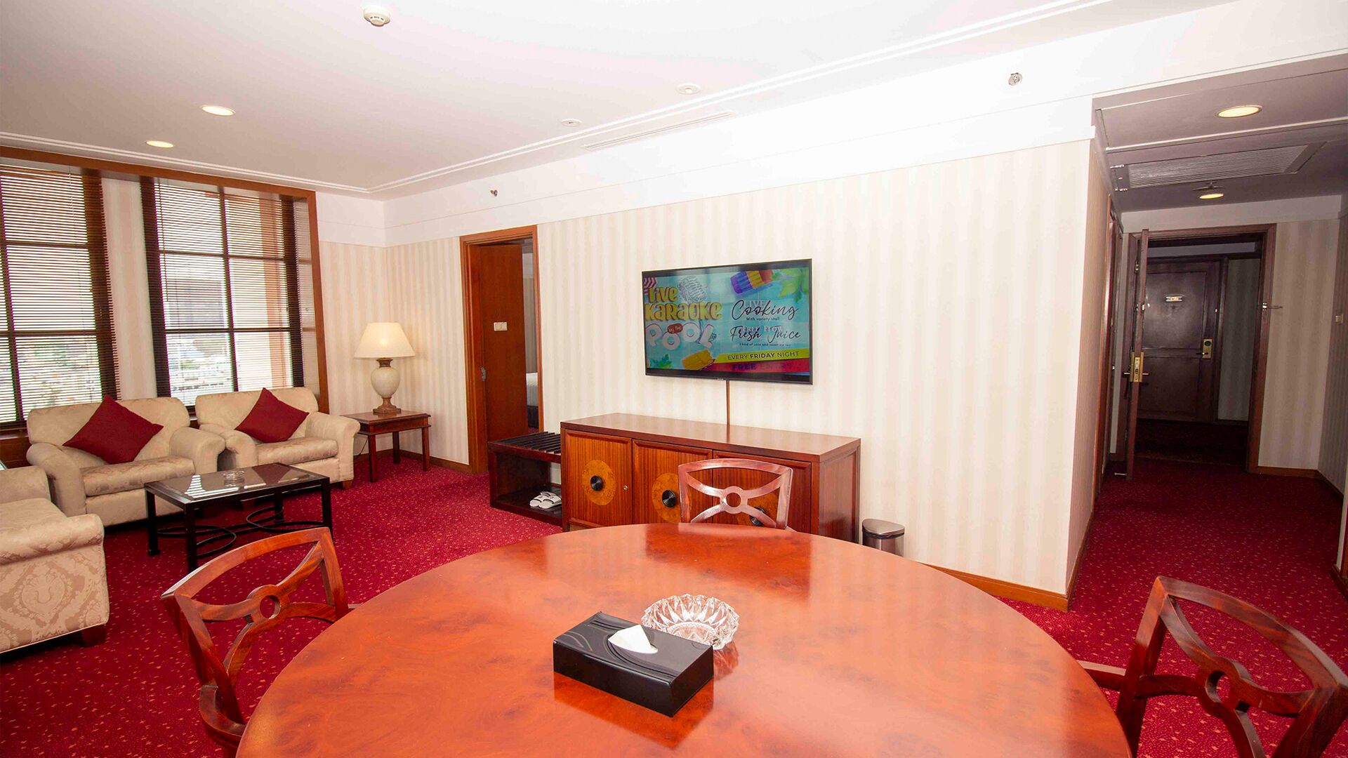 A hotel room with a red carpet, a wooden dining table, a sofa, and a television on the wall.